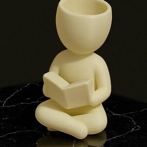 Puede incluir: Figura de color crema de una persona sentada con las piernas cruzadas leyendo un libro. La figura tiene una cabeza grande con la parte superior abierta, posiblemente para guardar objetos peque&ntilde;os. La figura est&aacute; sobre una superficie negra.