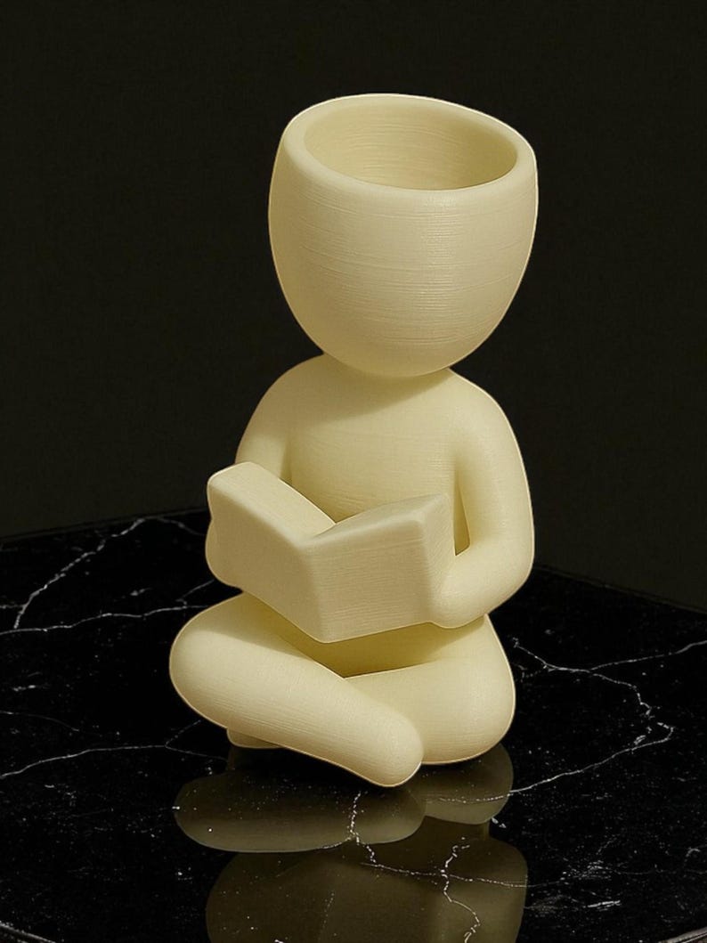 Puede incluir: Figura de color crema de una persona sentada con las piernas cruzadas leyendo un libro. La figura tiene una cabeza grande con la parte superior abierta, posiblemente para guardar objetos peque&ntilde;os. La figura est&aacute; sobre una superficie negra.