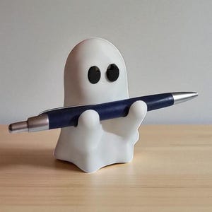 May include: A white ghost-shaped pen holder with black eyes, holding a blue and silver pen. The pen holder is on a light wooden surface, creating a playful and whimsical desk accessory.