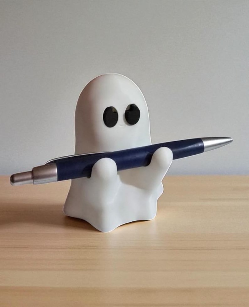 May include: A white ghost-shaped pen holder with black eyes, holding a blue and silver pen. The pen holder is on a light wooden surface, creating a playful and whimsical desk accessory.