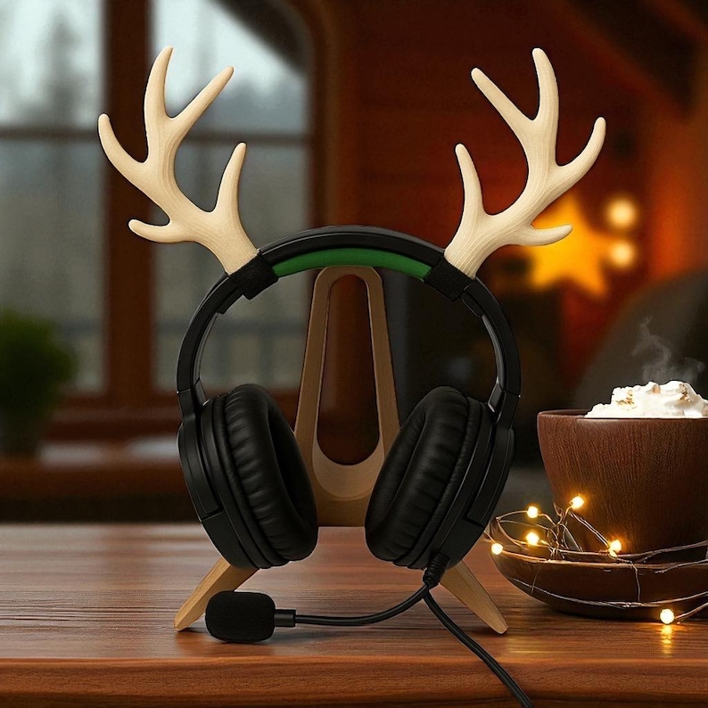 Op de afbeelding: Een houten hoofdtelefoonstandaard met rendiergewei, die een zwarte headset met een microfoon vasthoudt. De standaard staat op een houten tafel, met een kopje slagroom en lichtslingers op de achtergrond.