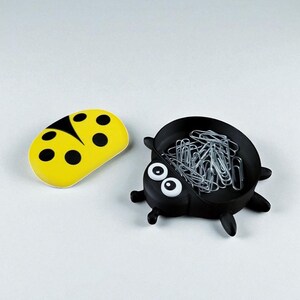 Puede incluir: Un sujetapapeles con forma de mariquita amarilla con lunares negros y un recipiente negro con forma de mariquita lleno de clips plateados. El recipiente tiene ojos blancos y peque&ntilde;as patas negras.