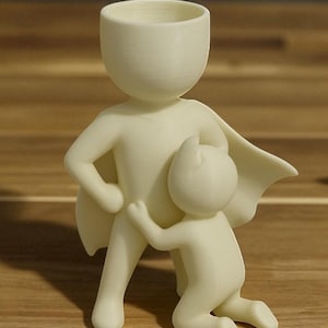 May include: A cream-colored figurine depicting a parent figure with a cape, embracing a smaller child figure. The parent figure has a cup-shaped head. The child figure is kneeling and hugging the parent's leg. The figures are on a wooden surface.