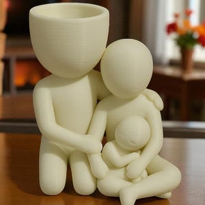 Puede incluir: Figura familiar de color crema, que representa a una figura paterna con una cabeza grande y una peque&ntilde;a abertura para maceta, abrazando a otro padre y a un beb&eacute;. Las figuras son estilizadas y lisas, con un acabado mate.