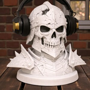 May include: A white, detailed skull bust wearing a helmet and armor, with black and gold headphones resting on its head. The bust is on a white circular base, set against a brick wall and wooden surface.
