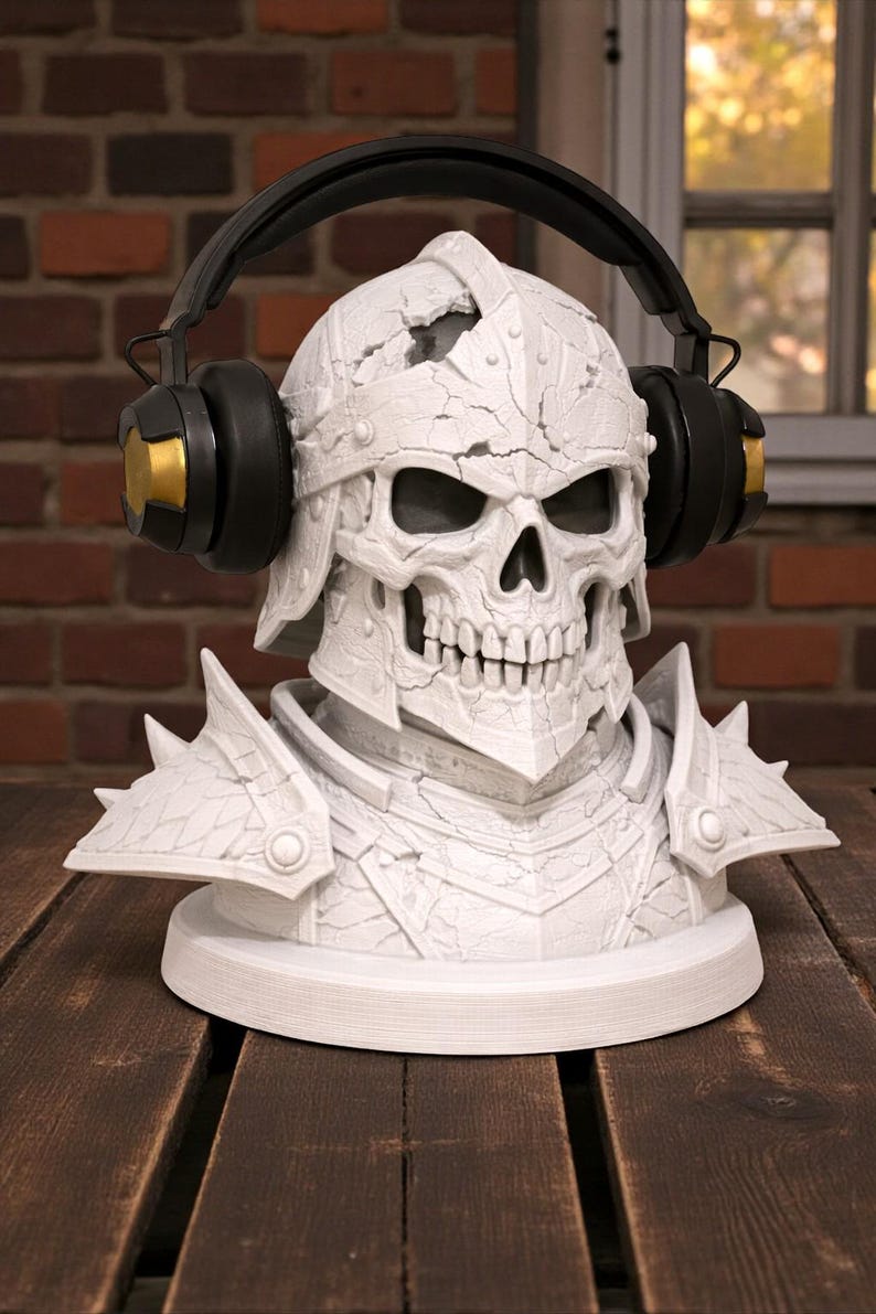 May include: A white, detailed skull bust wearing a helmet and armor, with black and gold headphones resting on its head. The bust is on a white circular base, set against a brick wall and wooden surface.