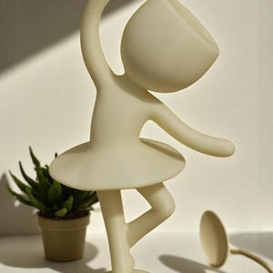 May include: A cream-colored figurine of a ballerina in a graceful pose, with one leg extended and arms outstretched. The figurine has a tutu and a rounded head. A small potted plant is in the background.