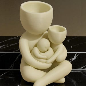 K&ouml;nnte beinhalten: Cremefarbene Skulptur, die eine sitzende Elternfigur mit zwei Kindern darstellt. Der Kopf des Elternteils hat die Form einer gro&szlig;en, offenen Schale. Die Kinder sind in den Armen des Elternteils eingebettet. Die Skulptur steht auf einer schwarz-wei&szlig;en Marmoroberfl&auml;che.