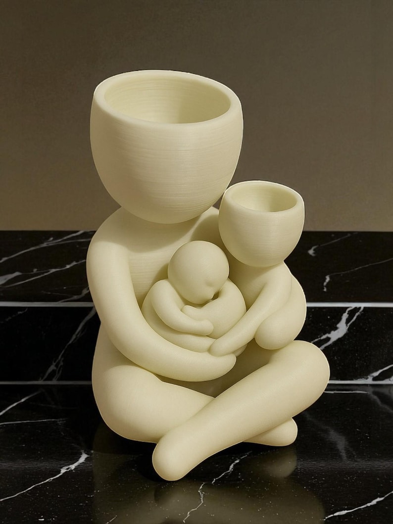 K&ouml;nnte beinhalten: Cremefarbene Skulptur, die eine sitzende Elternfigur mit zwei Kindern darstellt. Der Kopf des Elternteils hat die Form einer gro&szlig;en, offenen Schale. Die Kinder sind in den Armen des Elternteils eingebettet. Die Skulptur steht auf einer schwarz-wei&szlig;en Marmoroberfl&auml;che.