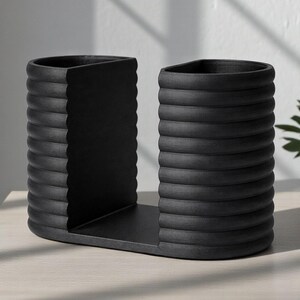 May include: A black, modern bookend set. The bookends have a unique design with a series of horizontal ridges, giving them a textured appearance. The base is a flat, rectangular shape, and the overall design is minimalist and stylish.