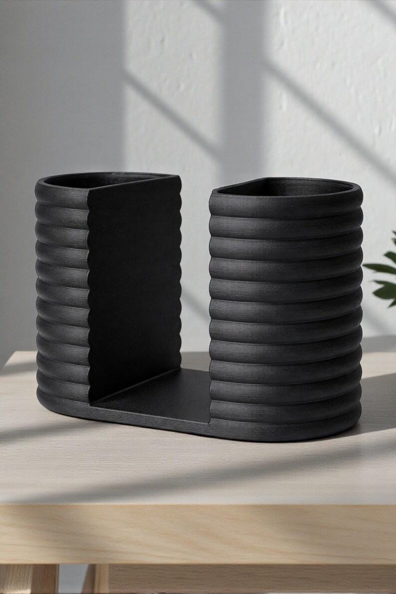 May include: A black, modern bookend set. The bookends have a unique design with a series of horizontal ridges, giving them a textured appearance. The base is a flat, rectangular shape, and the overall design is minimalist and stylish.