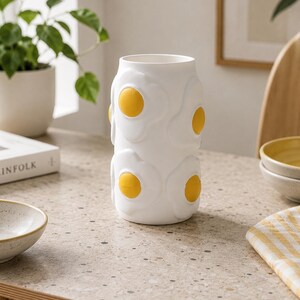 May include: A white ceramic vase with a repeating pattern of fried eggs, each with a yellow yolk. The vase is cylindrical and sits on a speckled countertop. The design is whimsical and adds a touch of fun to home decor.