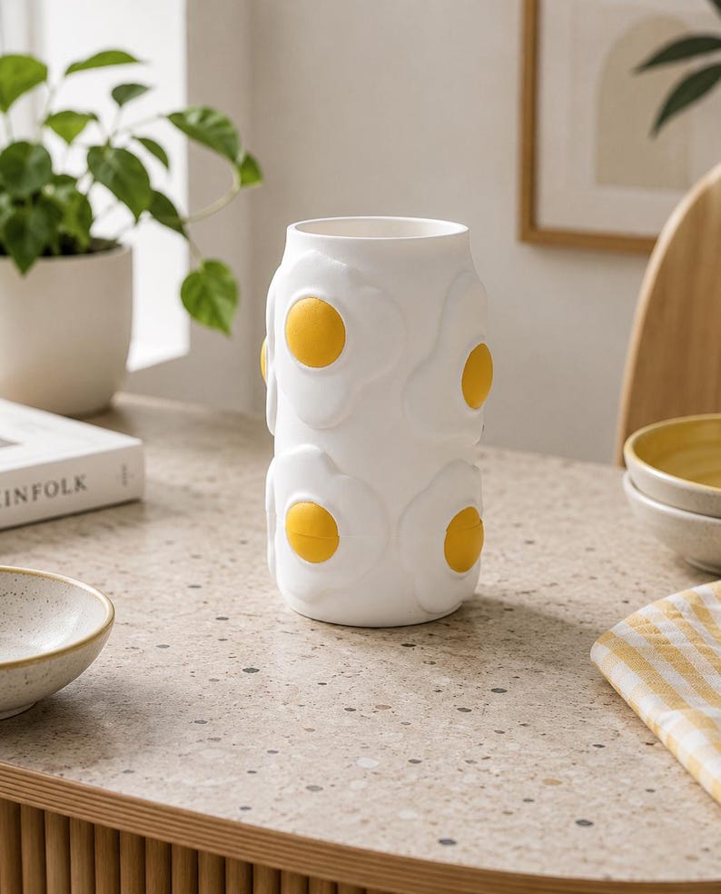 May include: A white ceramic vase with a repeating pattern of fried eggs, each with a yellow yolk. The vase is cylindrical and sits on a speckled countertop. The design is whimsical and adds a touch of fun to home decor.