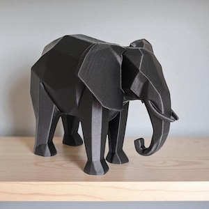 Op de afbeelding: Een zwarte geometrische olifant sculptuur met een facetontwerp. De olifant heeft grote oren, een slurf en vier poten. Hij staat op een lichtgekleurde houten plank tegen een grijze muur. De sculptuur is ongeveer 25 cm lang.