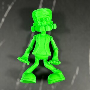 May include: A green plastic figurine of Frankenstein's monster with a goofy expression. The figurine has a textured surface and is posed with its arms outstretched and legs bent.