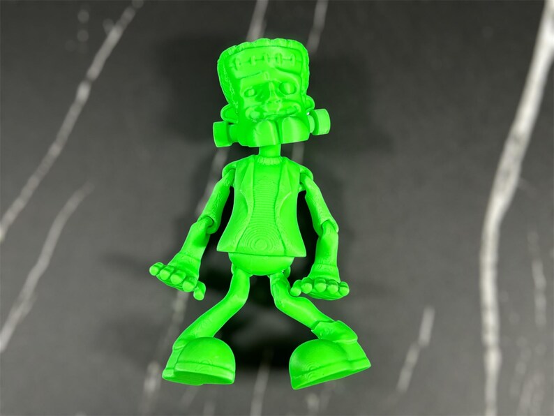 May include: A green plastic figurine of Frankenstein's monster with a goofy expression. The figurine has a textured surface and is posed with its arms outstretched and legs bent.