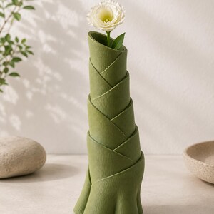 May include: A green, bamboo-shaped vase holding a white flower with a green stem and leaf. The vase has a unique, layered design and sits on a light-colored surface. The vase is approximately 10 inches tall.