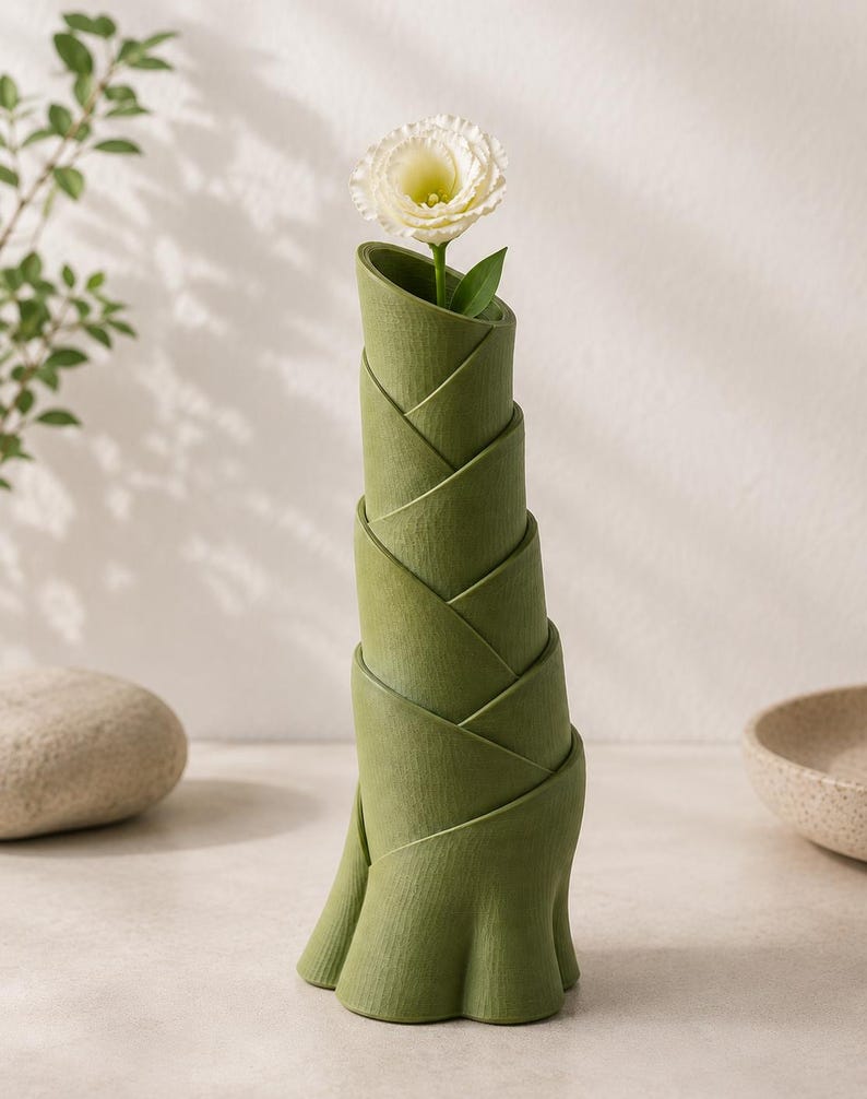 May include: A green, bamboo-shaped vase holding a white flower with a green stem and leaf. The vase has a unique, layered design and sits on a light-colored surface. The vase is approximately 10 inches tall.