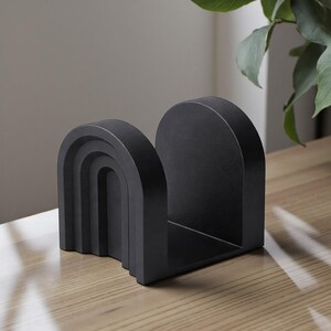 以下が含まれることがあります： 黒いアーチ型のブックエンド。層状の虹のデザイン。ブックエンドは無垢材でできており、明るい色の木製の表面に置かれています。デザインはモダンでミニマリストです。