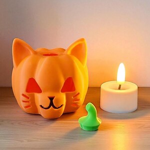 May include: Orange cat-shaped Halloween candle holder with cut-out features. A small green decorative piece and a white candle with a lit flame are next to the cat. The items are on a wooden surface.