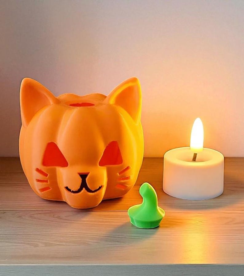 May include: Orange cat-shaped Halloween candle holder with cut-out features. A small green decorative piece and a white candle with a lit flame are next to the cat. The items are on a wooden surface.