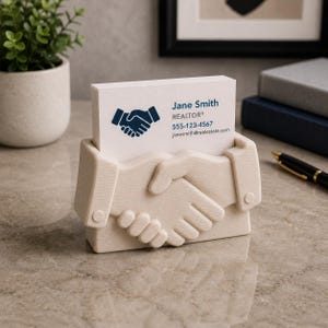 May include: A beige business card holder shaped like two hands clasped in a handshake. The holder displays a stack of white business cards with the text "Jane Smith, REALTOR&reg;" and contact information. The design is a symbol of agreement.