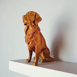 Op de afbeelding: Een goudkleurig hondenbeeld in zittende positie. De sculptuur heeft gedetailleerde vacht- en gezichtskenmerken en staat op een witte plank tegen een effen witte muur. De hond kijkt naar links.