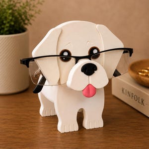 May include: A white dog-shaped eyeglasses holder with black-framed glasses. The dog has a black nose, brown eyes, and a pink tongue. The holder is made of wood and sits on a wooden surface.
