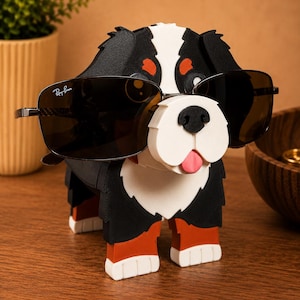 May include: A black, white, and red dog-shaped sunglasses holder with a pair of Ray-Ban sunglasses. The dog has a white muzzle, a pink tongue, and orange accents. The sunglasses are black with silver frames.