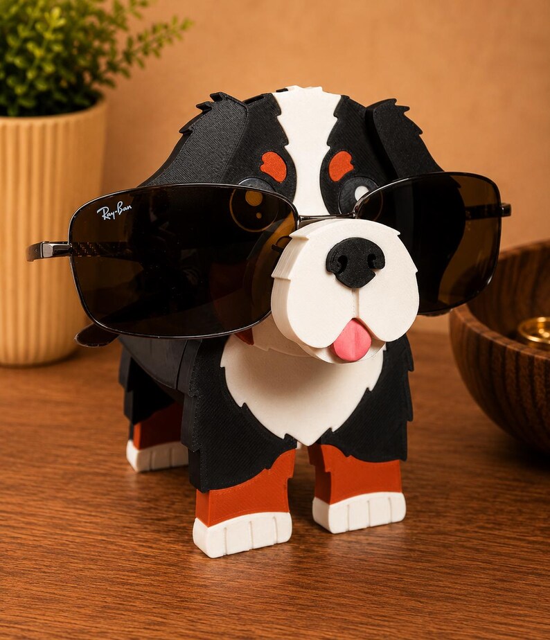 May include: A black, white, and red dog-shaped sunglasses holder with a pair of Ray-Ban sunglasses. The dog has a white muzzle, a pink tongue, and orange accents. The sunglasses are black with silver frames.