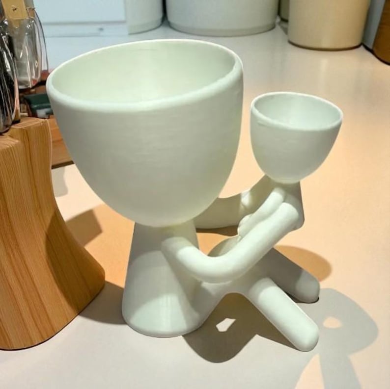 Op de afbeelding: Een witte keramische plantenbak in de vorm van een persoon die een kleinere plantenbak vasthoudt. De grotere plantenbak heeft een brede, open komvorm, terwijl de kleinere in de armen van de figuur wordt gehouden. Het ontwerp is minimalistisch en modern.