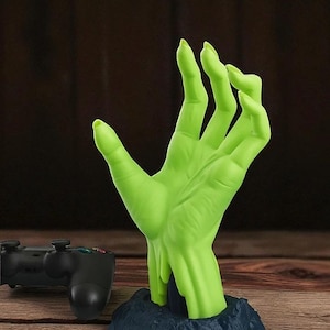 Pode incluir: Uma mão verde estilizada emergindo de uma base azul escura. A mão tem dedos longos e pontudos e uma superfície texturizada. Um controle de jogo preto é visível no fundo, sugerindo um item com tema de jogos.