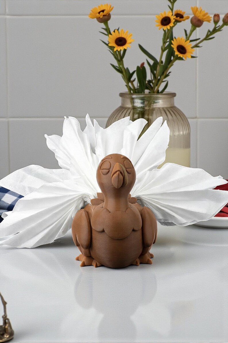 Puede incluir: Un servilletero marr&oacute;n con forma de pavo, con un dise&ntilde;o detallado, sostiene una servilleta blanca. El pavo est&aacute; sobre una superficie blanca. Un jarr&oacute;n con flores amarillas est&aacute; en el fondo.