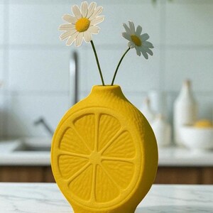 May include: A bright yellow ceramic vase shaped like a lemon slice, holding two daisies. The vase has a textured surface and is set on a white countertop. The daisies have white petals and yellow centers. The background includes a kitchen sink and other decor.