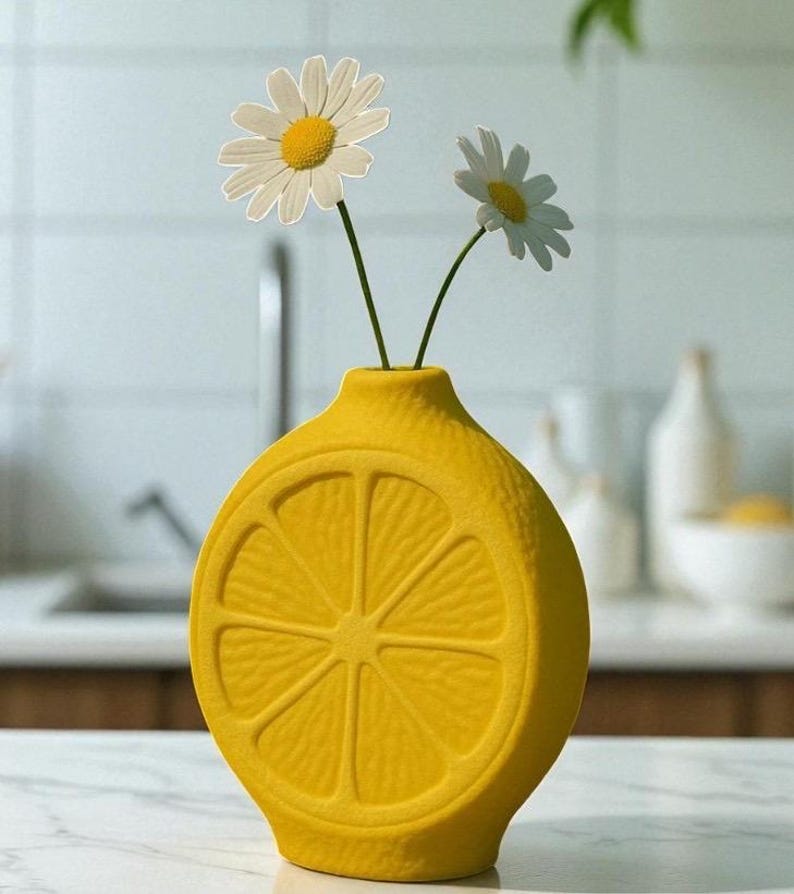 May include: A bright yellow ceramic vase shaped like a lemon slice, holding two daisies. The vase has a textured surface and is set on a white countertop. The daisies have white petals and yellow centers. The background includes a kitchen sink and other decor.