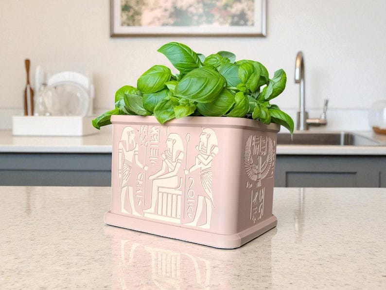 May include: A pink rectangular planter with white Egyptian hieroglyphs and a base, filled with vibrant green basil. The planter is on a countertop in a kitchen setting. The design features pharaohs and other ancient Egyptian symbols.