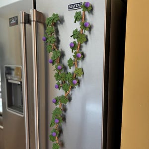 May include: A decorative vine with green leaves and purple berries is attached to a stainless steel refrigerator. The vine is a whimsical accent, adding a touch of nature to the kitchen decor. The refrigerator has a Maytag logo.