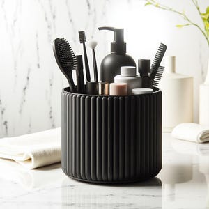 May include: A black, ribbed cylindrical container holding various bathroom accessories. Items include a hairbrush, toothbrush, makeup brushes, and a soap dispenser. The container sits on a white marble surface.