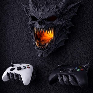 May include: A dark gray demonic skull with glowing orange light emanating from its mouth, above two game controllers. One controller is white, the other black. The controllers are held by clawed hands.