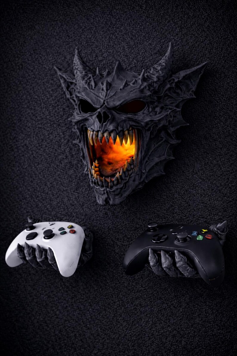 May include: A dark gray demonic skull with glowing orange light emanating from its mouth, above two game controllers. One controller is white, the other black. The controllers are held by clawed hands.