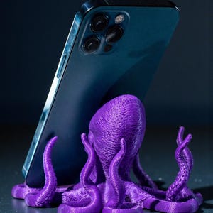 May include: A purple octopus-shaped phone stand supporting a blue smartphone. The stand is 3D-printed and features detailed tentacles and a textured body. The phone is angled against the octopus, creating a unique desk accessory.