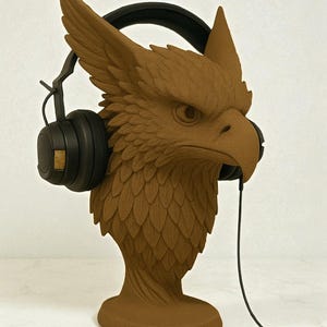 May include: A brown griffin head sculpture wearing black headphones. The sculpture has detailed feather-like textures and is mounted on a pedestal base. The headphones have black earcups and a black headband.