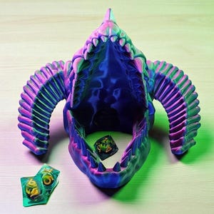 Op de afbeelding: Een veelkleurige, 3D-geprinte drakenschedel met een open mond. De schedel is voornamelijk blauw, met roze en groene accenten. Twee dobbelstenen liggen in de mond, en twee dobbelstenen liggen ernaast.