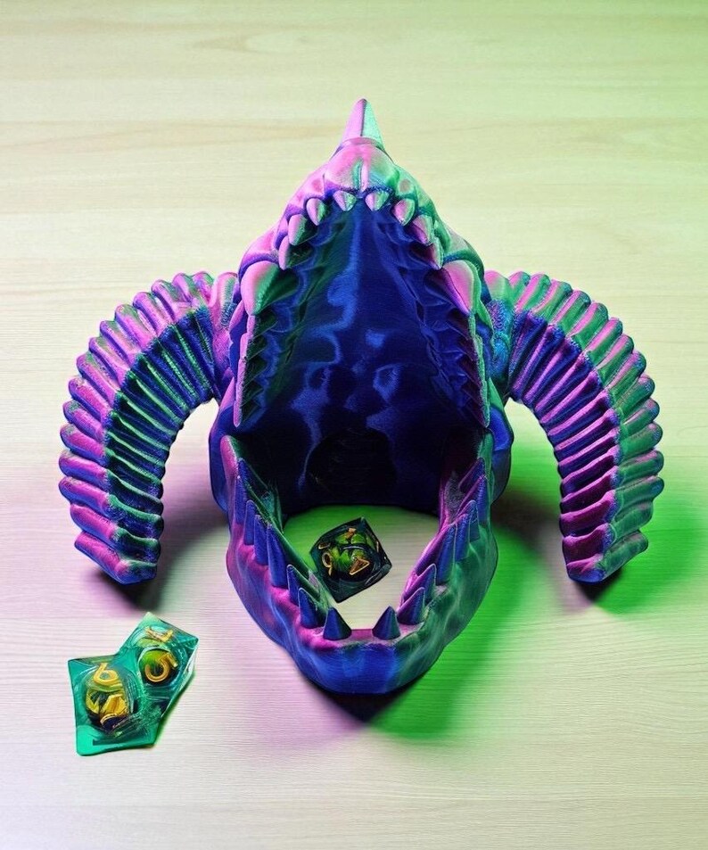 Op de afbeelding: Een veelkleurige, 3D-geprinte drakenschedel met een open mond. De schedel is voornamelijk blauw, met roze en groene accenten. Twee dobbelstenen liggen in de mond, en twee dobbelstenen liggen ernaast.