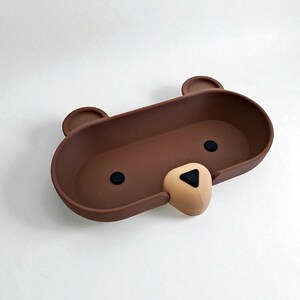 Op de afbeelding: Een bruine, beer-vormige tray met een afgerond rechthoekig ontwerp. De tray heeft twee kleine, afgeronde oren, twee zwarte cirkelvormige ogen en een bruine neus met een zwarte driehoek. De tray is ontworpen voor het vasthouden van kleine items.