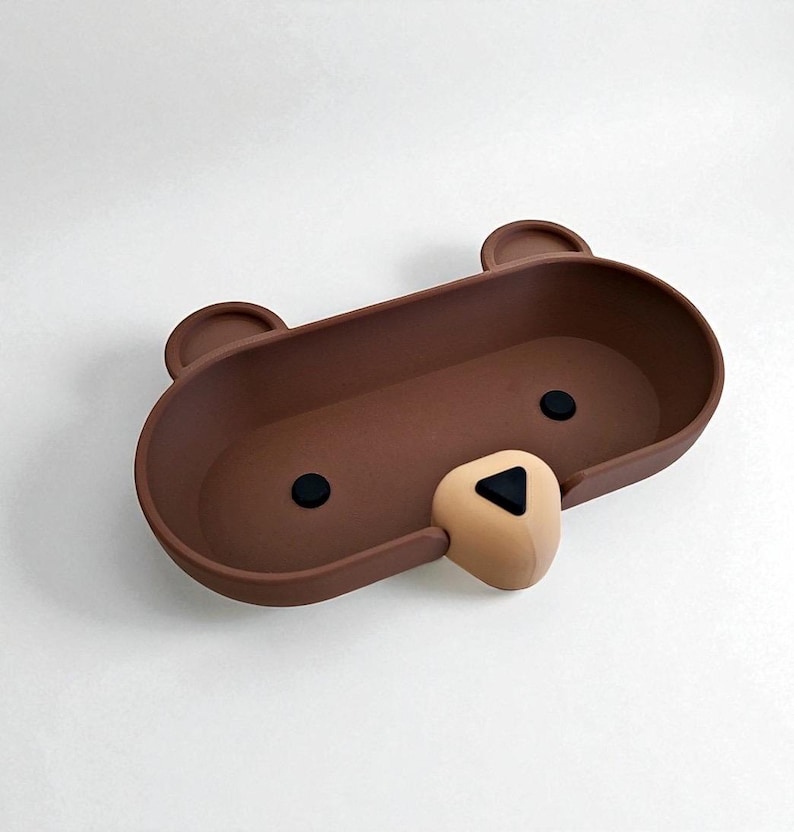 Op de afbeelding: Een bruine, beer-vormige tray met een afgerond rechthoekig ontwerp. De tray heeft twee kleine, afgeronde oren, twee zwarte cirkelvormige ogen en een bruine neus met een zwarte driehoek. De tray is ontworpen voor het vasthouden van kleine items.