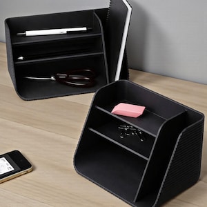 May include: Two black desk organizers with multiple compartments. One contains a white pen, a pencil, and scissors. The other holds a pink eraser and paper clips. A smartphone rests on the wooden surface.