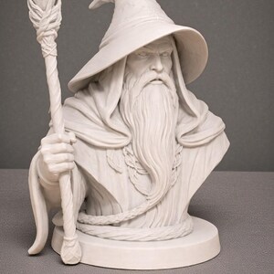 May include: A white bust of a wizard with a long beard, a pointed hat, and a staff topped with a crystal. The wizard's face has a stern expression. The bust is on a round base.