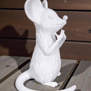 May include: A white ceramic mouse figurine with a textured surface. The mouse is standing upright with its hands near its face and a long, curved tail. The figurine has large ears and a small nose. The background is a wooden surface.