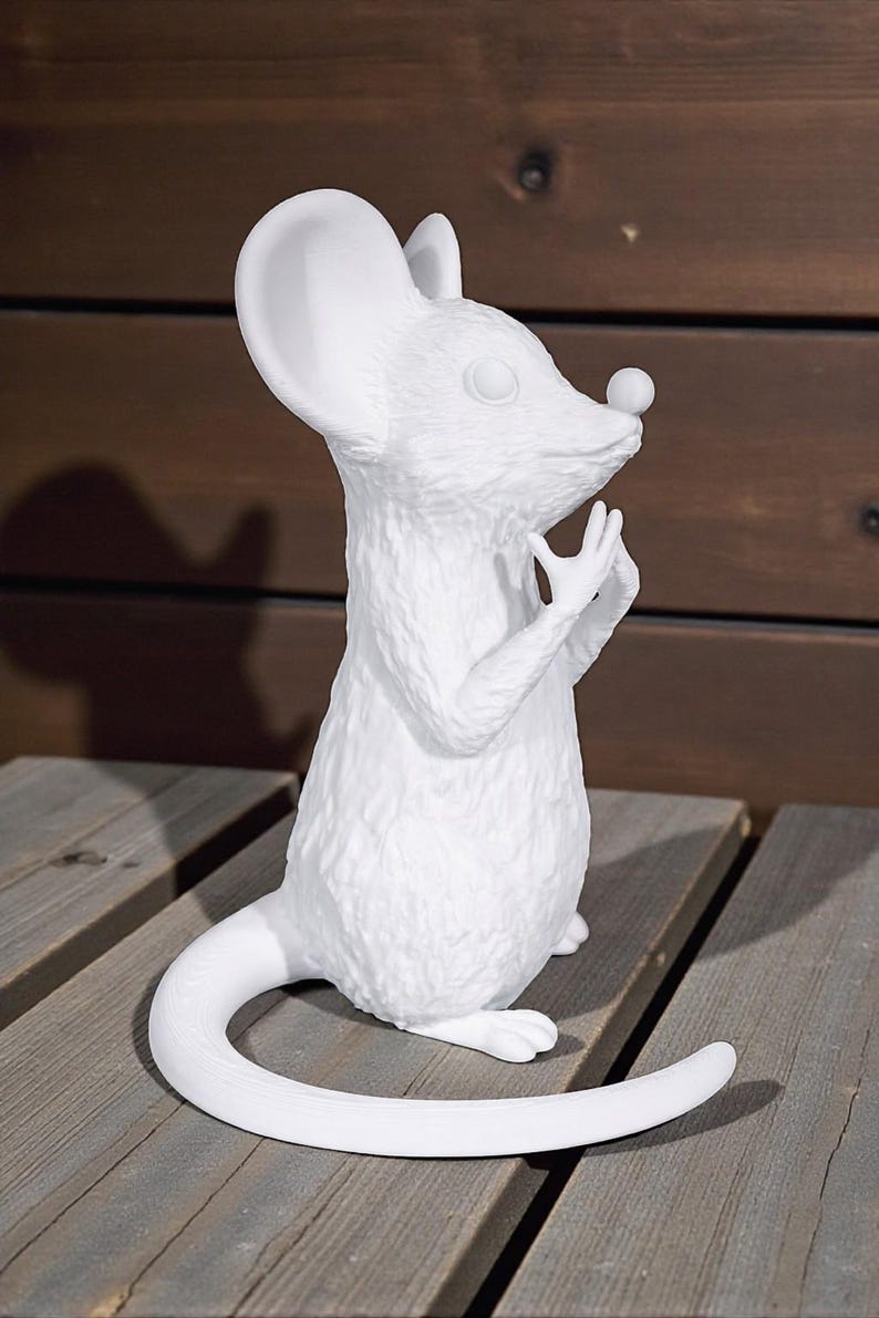 May include: A white ceramic mouse figurine with a textured surface. The mouse is standing upright with its hands near its face and a long, curved tail. The figurine has large ears and a small nose. The background is a wooden surface.